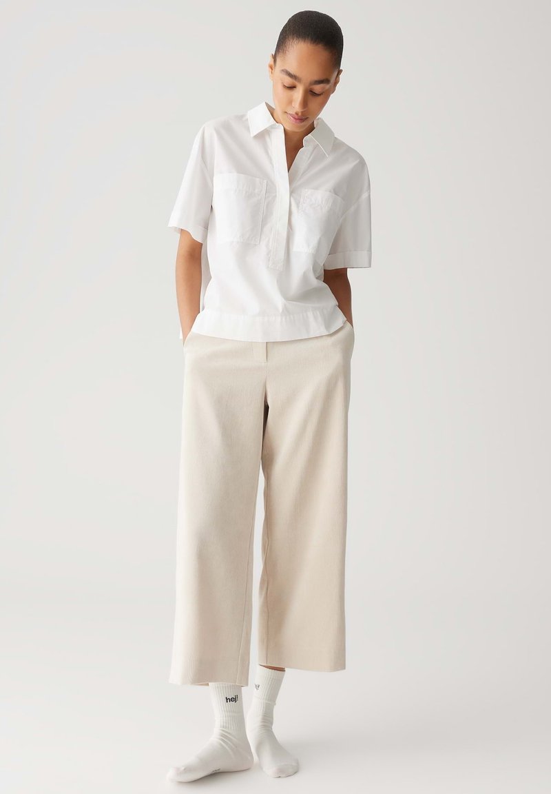 Weißes Kurzarmhemd mit zwei Brusttaschen, kombiniert mit beige-weißen weiten Hosen. Das Modell trägt weiße Socken mit Schriftzug. Einfaches, sauberes Design.