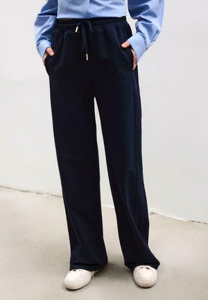 Person iført sorte, vidt benbukser med snøre i taljen, lyseblå langærmet skjorte og hvide sneakers, stående mod en ensfarvet væg.