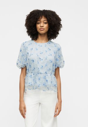 Femme aux cheveux bouclés portant un chemisier léger bleu clair avec broderie florale bleue et un pantalon blanc, debout devant un fond uni.