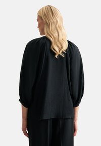 Blusa nera con vestibilità rilassata, caratterizzata da maniche a sbuffo, tessuto morbido e dettaglio con bottone sui polsini. Taglio leggermente più lungo sul retro.
