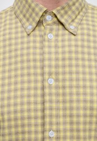 Chemise à carreaux jaune et beige avec boutons, col boutonné et boutons blancs attachés au niveau du col.