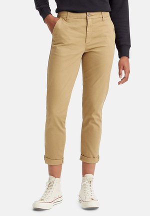 Person iført beige op-rullede chinos, sort langærmet top og hvide høje sneakers, står med den ene hånd i lommen.
