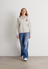 Grå fleece-pullover med krage och knappar, prydd med ett rött bokstav "G" på bröstet, i kombination med ljusblå vida jeans och vita skor.
