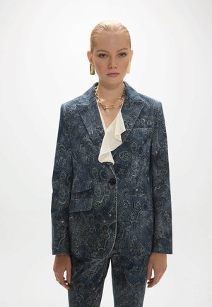 Mujer con traje oscuro de estampado paisley, blusa blanca con volantes, collar de cadena dorado y pendientes rectangulares dorados sobre fondo blanco.