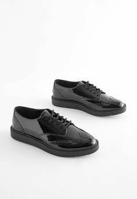 Čierne lakované šnurovacie topánky s brogue detailmi, s hladkým, lesklým povrchom, zaoblenou špičkou a hrubou, plochou gumovou podrážkou.