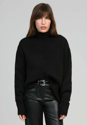 Femme aux cheveux bruns portant un pull à col roulé noir et un pantalon en cuir noir taille haute avec une ceinture, debout devant un fond neutre.