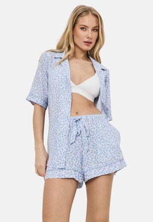 Vrouw draagt lichtblauw gebloemd kort mouws knoophemd en bijpassende korte broek met witte bralette tegen een witte achtergrond.