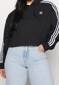 Hoodie cropped nero con design a tre strisce bianche sulle maniche e piccolo logo Adidas bianco sul petto. Indossato con jeans chiari a vita alta.