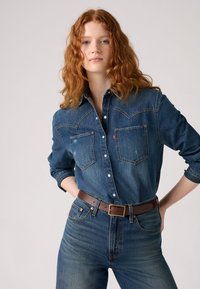 Kvinne med langt rødt krøllete hår som har på seg en knappet denimskjorte stukket ned i høyhalsede denimjeans med et brunt belte.