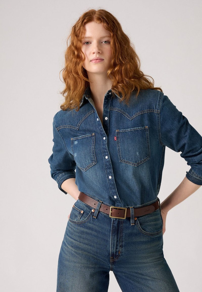 Kvinne med langt rødt krøllete hår som har på seg en knappet denimskjorte stukket ned i høyhalsede denimjeans med et brunt belte.