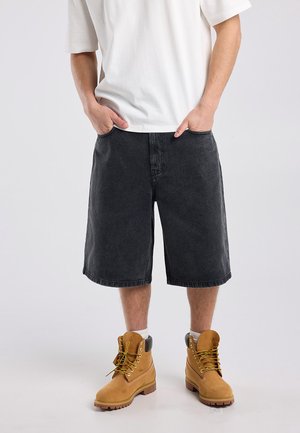 Man wearing white t-shirt, long black denim shorts, white socks, and tan lace-up work boots standing with hands in pockets.