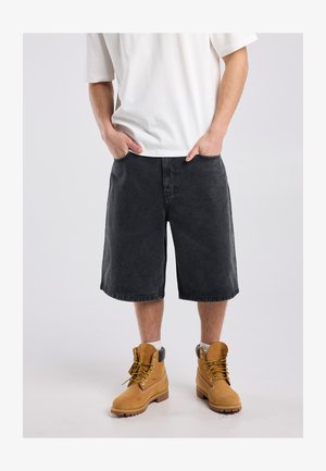 Man draagt een wit T-shirt, lange zwarte denimshorts, witte sokken en beige veterwerklaarzen, staat met handen in de zakken.