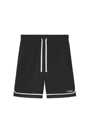 Persona con pantalones cortos de malla negros con ribete blanco, calcetines blancos con texto negro y mocasines en blanco y negro, de pie contra un fondo liso.