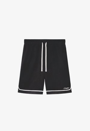 Person trägt schwarze Netzshorts mit weißem Saum, weiße Socken mit schwarzem Text und schwarz-weiße Loafer und steht vor einem einfarbigen Hintergrund.
