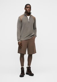 Άνδρας που φοράει μπεζ πουλόβερ με φερμουάρ έως τη μέση, καφέ βερμούδα μέχρι το γόνατο, λευκές κάλτσες και σκούρα καφέ δερμάτινα παπούτσια, στέκεται μπροστά σε απλό φόντο.