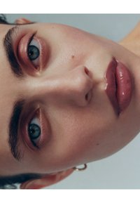 Lähikuva kasvoista, joissa on kirkas iho, siniset silmät, selkeästi muotoillut kulmakarvat ja kiiltävät vaaleanpunaiset huulet. Hillitty poskipuna ja pieni korvakoru somisteena.