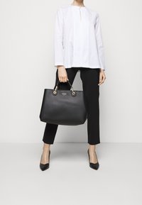 Sac en cuir noir avec quincaillerie en ton doré et deux poignées supérieures. Surface texturée sans motifs visibles, associé à un haut blanc et un pantalon noir.