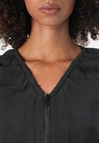 Svart zip-up topp med V-hals, med en samlet tekstur rundt halsen og et glatt, lett stoff.