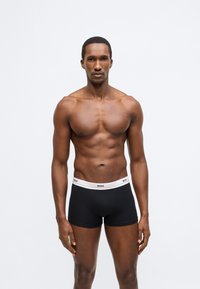 Boxeri negri cu talie albă pe care este imprimat logo-ul "BOSS". Purtați de un model cu o constituție atletică. Design simplu, fără modele vizibile.
