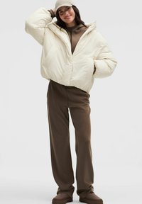 Skračená puffová bunda v odtieňoch off-white, vybavená kapucňou a dvoma prednými vreckami, skombinovaná s hnedými nohavicami a ladiacimi slip-on topánkami.