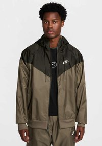 Kurtka Nike typu zip-up w jasno-oliwkowym i ciemno-oliwkowym kolorze. Posiada kaptur, elastyczne mankiety oraz wykończenie. Logo na piersi. Gładki, lekki materiał.