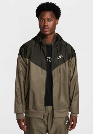 Giacca zip-up Nike in verde oliva chiaro e scuro. Caratterizzata da un cappuccio, polsini ed orlo elasticizzati. Logo sul petto. Materiale morbido e leggero.