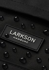 Svart tyg med vattendroppar, med en svart och vit etikett som läser "LARKSON HAMBURG" fäst vid materialet.