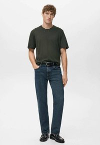 Ciemnozielona bawełniana koszulka o luźnym kroju, zestawiona z niebieskimi dżinsami o prostych nogawkach, dopełniona czarnymi skórzanymi butami.