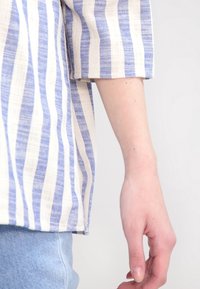 Chemise rayée avec des lignes verticales bleues et crème, fabriquée en tissu texturé. Manches courtes avec une coupe décontractée, associée à un denim bleu clair.