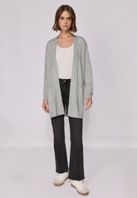 Světle šedý cardigan vyrobený z měkké tkaniny, otevřený design s dlouhými rukávy, kombinovaný s bílým topem, černými rozšířenými džíny a bílými teniskami.