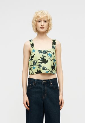 Fiatal személy göndör szőke hajjal, zöld ujjatlan haspólót visel, amelyen fekete madár és kék csillag minták vannak, valamint sötét farmernadrágot.