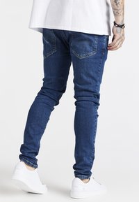 Slim-fit blue jeans with a smooth denim texture, five-pocket design, and subtle stitching details, paired with white sneakers.