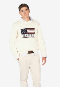 Jersey de punto color crema con un diseño de bandera en azul y rojo y la palabra "HARPER" en azul. Lleva puesto sobre una camisa con cuello y pantalones beige.