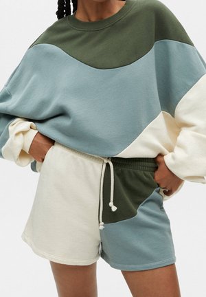 Personne portant un sweat-shirt color-block et un short assorti en vert olive, bleu clair et crème, les mains dans les poches de la ceinture.