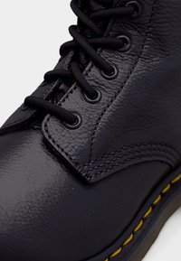 Botte en cuir noir à lacets avec surface texturée, œillets métalliques, coutures visibles et bord de semelle accentué en jaune.
