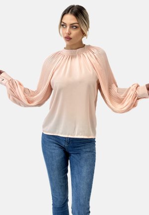 Blusa pesca chiaro con maniche plissettate, scollatura rotonda e polsini. Abbinata a jeans blu, caratterizzati da una superficie liscia e leggermente riflettente.