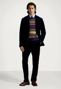 Άνδρας με σκούρο βελούδινο κοστούμι, πολύχρωμο μοτίβο πουλόβερ, μπλε πουκάμισο, ναυτικό λ tie και καφέ δερμάτινες λαστιχένιες μπότες, στέκεται με το ένα χέρι στην τσέπη.