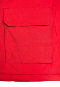 Rød stof med en tekstur og glans, der har en rektangulær patch-lomme med en klap i den øverste kant, syet med matchende tråd.