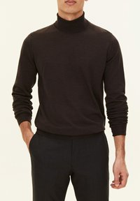 Uomo che indossa un maglione a collo alto marrone scuro a maniche lunghe con pantaloni neri, una mano in tasca, in piedi contro uno sfondo chiaro.