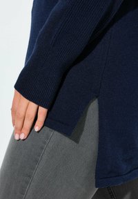 Marinblå stickad tröja med ribbade ärmar, delad fåll och mjukt material, kombinerad med grå jeans. Fokusera på tröjans detaljer och tyg.