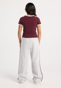 Persona in piedi con la schiena rivolta, indossa una maglietta attillata bordeaux con rifiniture bianche e pantaloni da jogging grigi larghi con strisce laterali.