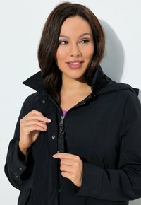 Femme aux cheveux bruns mi-longs portant une veste noire à col relevé, tenant la tirette de la fermeture éclair tout en regardant sur le côté.