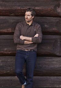 Mand med krydsede arme, der læner sig mod en mørk væg af træstammer, iført brun langærmet polo og blå jeans, kigger mod sin venstre side.