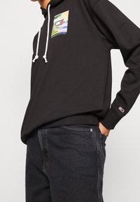 Felpa nera con cappuccio e lacci bianchi, caratterizzata da una stampa grafica multicolore sul petto. Abbinata a jeans in denim scuri.