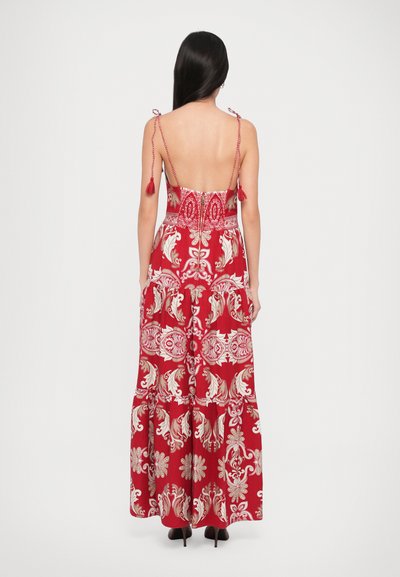 Red maxi dress with intricate white floral patterns, open back, and adjustable tie straps with tassels. Flowy fabric and tiered design.