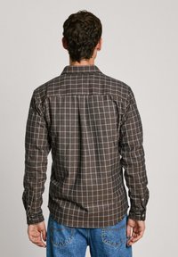 Bruine geruite shirt met een wit rasterpatroon, knopenmanchetten en een rugpaneel. Combineert goed met blauwe spijkerbroek, waarbij de gestructureerde stof zichtbaar is.