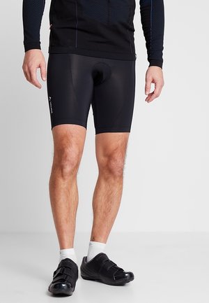Mann trägt schwarze Radhose, schwarze Radschuhe mit weißen Socken und ein schwarzes Langarmshirt, steht auf grauem Boden.