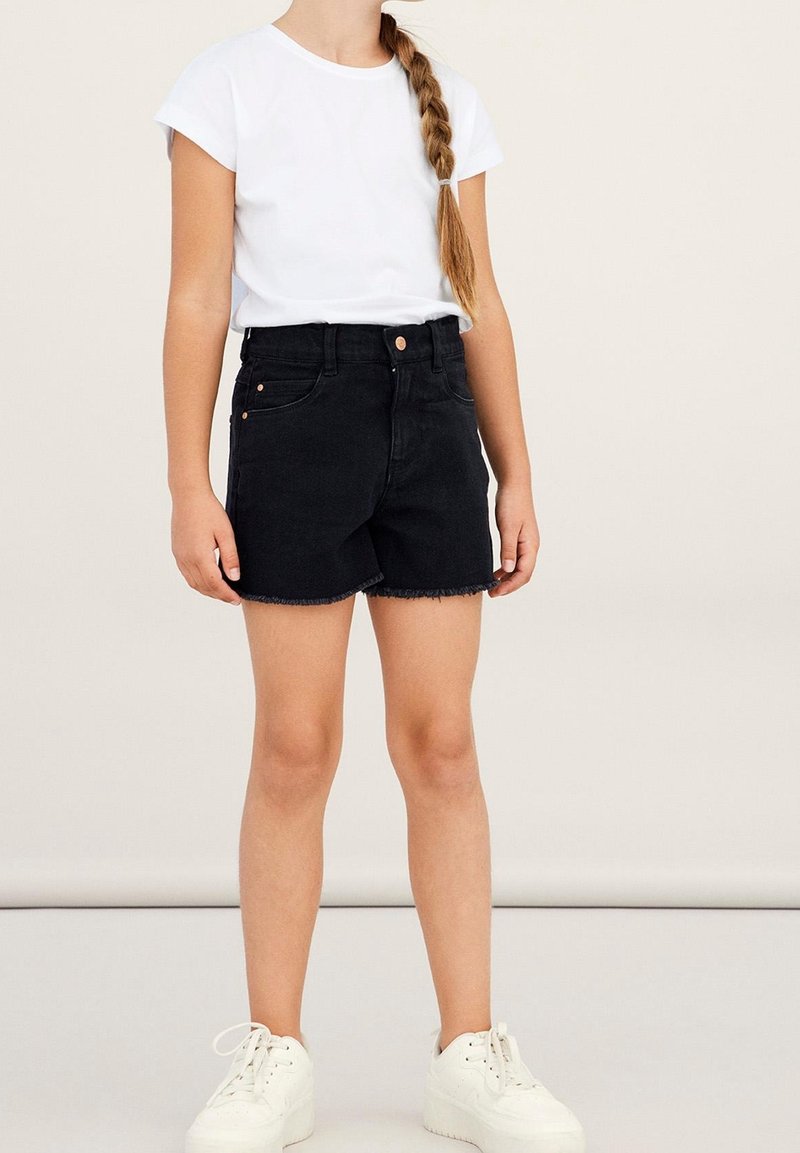 Jeune fille avec une longue tresse portant un t-shirt blanc, un short en jean noir et des baskets blanches, debout devant un fond clair uni.