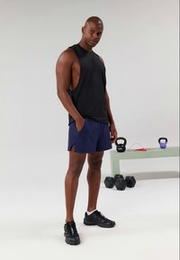 Mand i sort ærmeløs skjorte og marineblå shorts står ved en bænk med kettlebells, håndvægte og springreb i en minimal gym-opsætning.