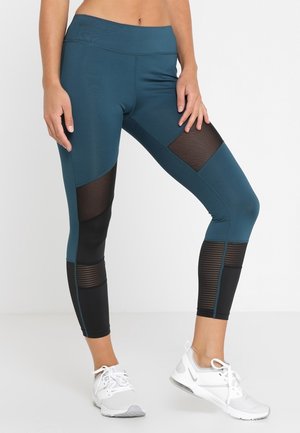 Femme portant des leggings avec des panneaux en mesh bleu sarcelle et noir et des chaussures de sport blanches, debout devant un fond clair uni.